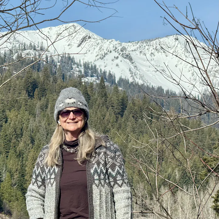 Portrait of Joan Spriggs, BCLD board member, smiling and standing in the outdoors wearing sweater, knit cap, and sunglasses, with woodland and snowy mountain range scenery.