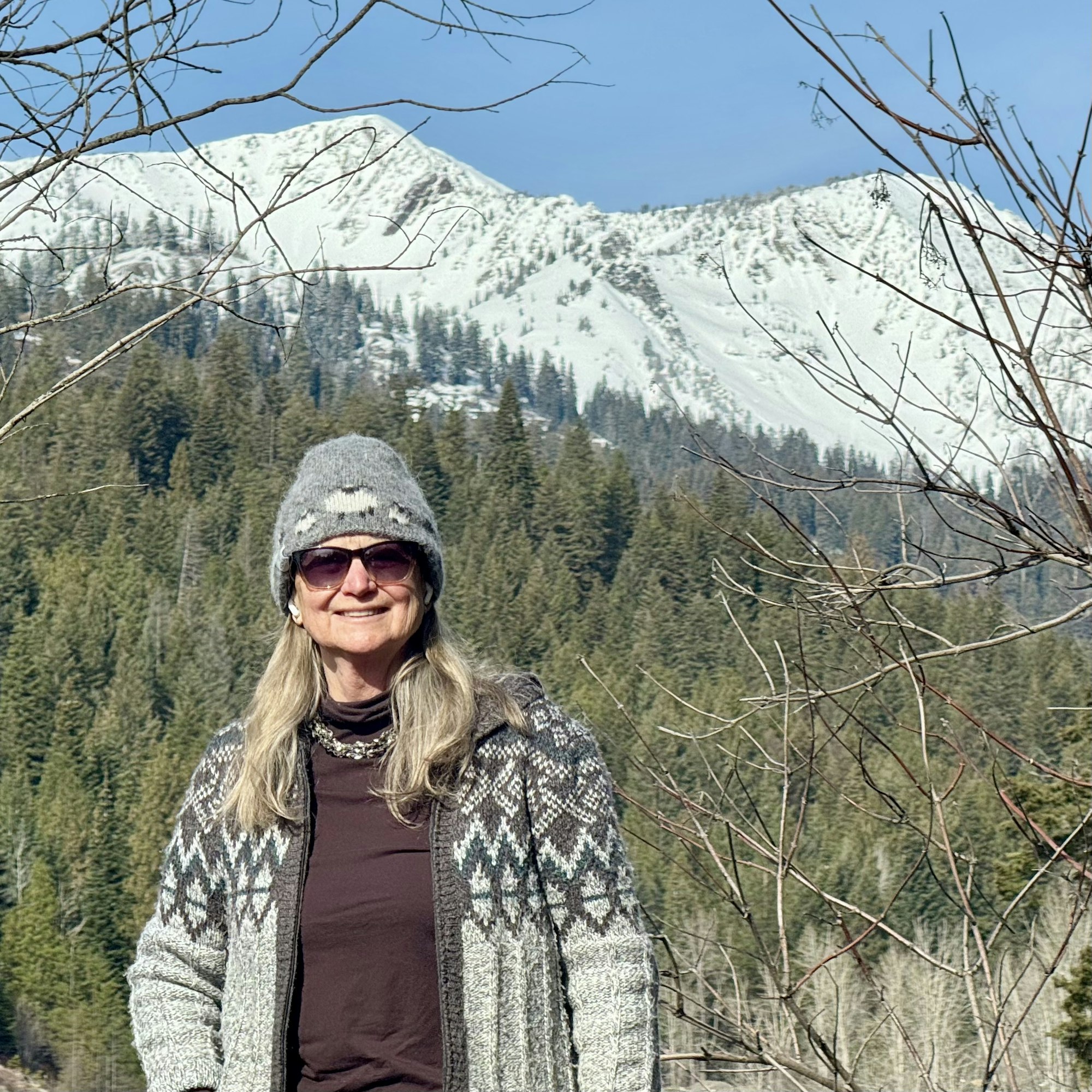Portrait of Joan Spriggs, BCLD board member, smiling and standing in the outdoors wearing sweater, knit cap, and sunglasses, with woodland and snowy mountain range scenery.