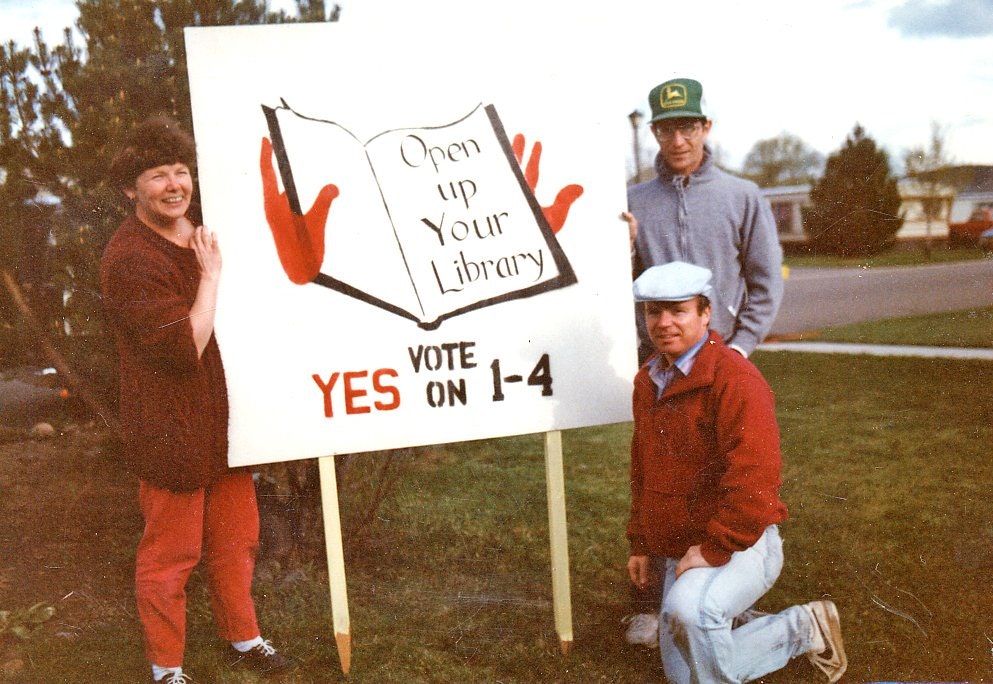 Three people promoting the library district initiative with a sign that says "Open up Your Library" and encourages voting on the proposal.