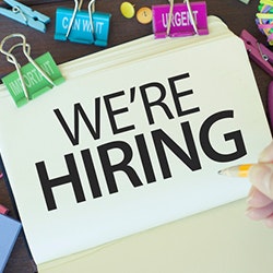 A "We're Hiring" sign on a desk with colorful clips, a person's hand visible.
