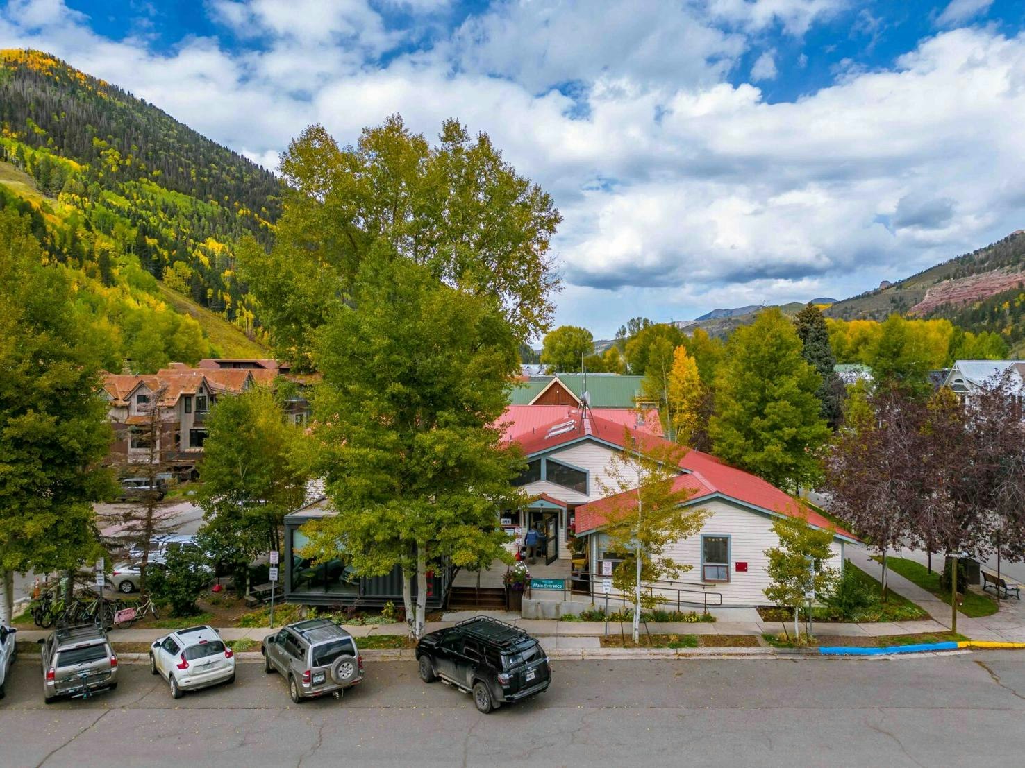 A picturesque scene featuring a quaint building, lined with cars, surrounded by colorful autumn trees and mountains under a cloudy sky.