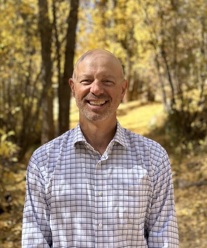 A person smiling in a checkered shirt stands in a wooded area with autumn foliage.