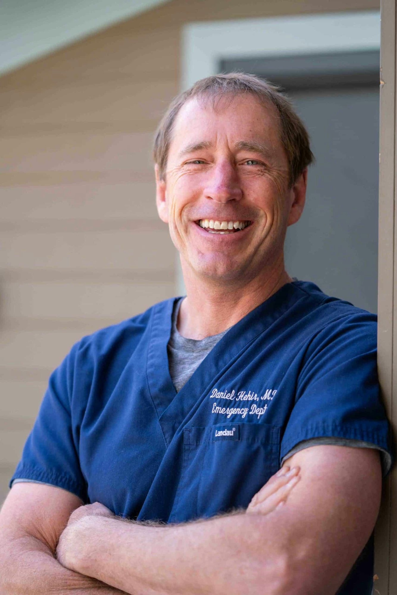 A smiling person in blue medical scrubs stands with arms crossed outdoors.
