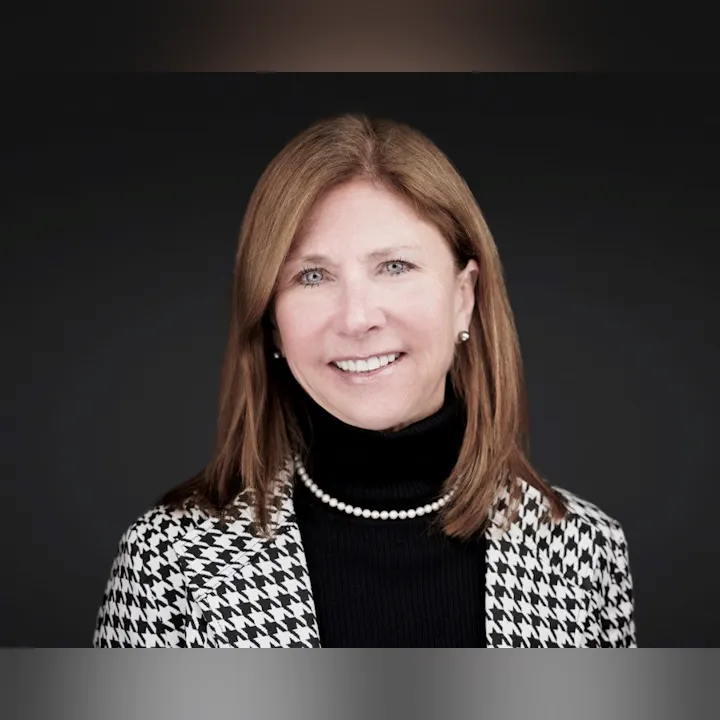A person with brown hair wearing a houndstooth jacket, black turtleneck, and pearl necklace, smiling against a dark background.