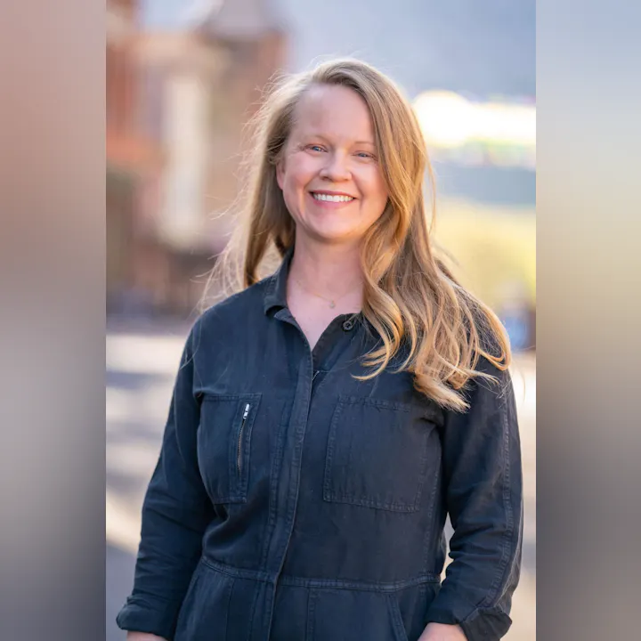 A person smiling outdoors, wearing a dark jacket.
