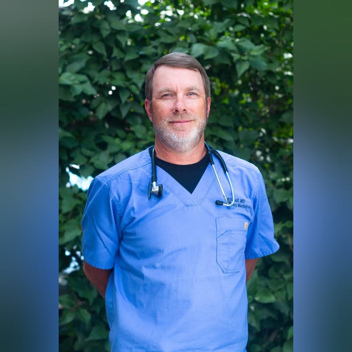 A person in blue scrubs with a stethoscope, standing in front of lush green foliage.