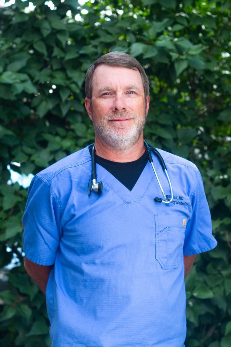 A person in blue scrubs with a stethoscope, standing in front of lush green foliage.