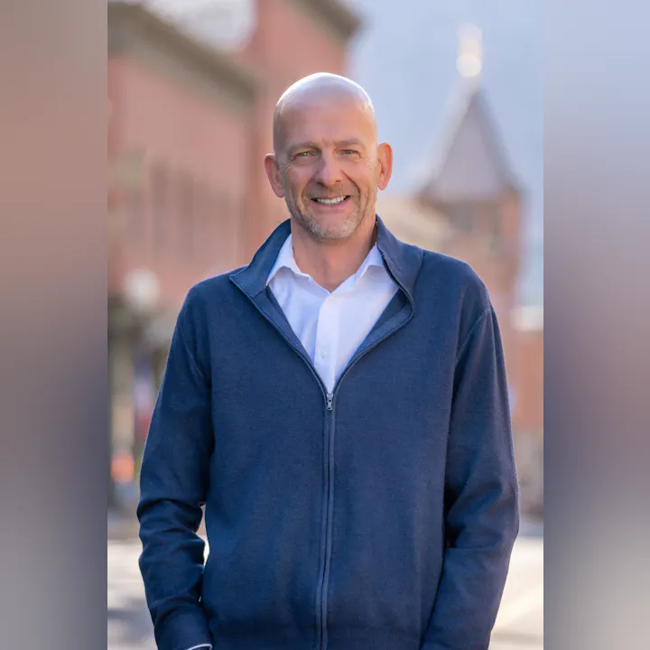 A person in a blue jacket smiles outdoors with a blurred building in the background.