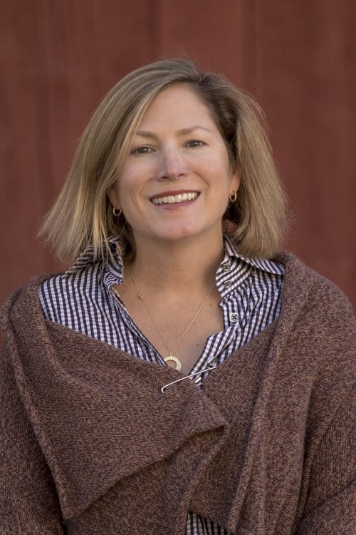 A smiling person with short hair, wearing a brown shawl over a checkered shirt, standing in front of a red background.