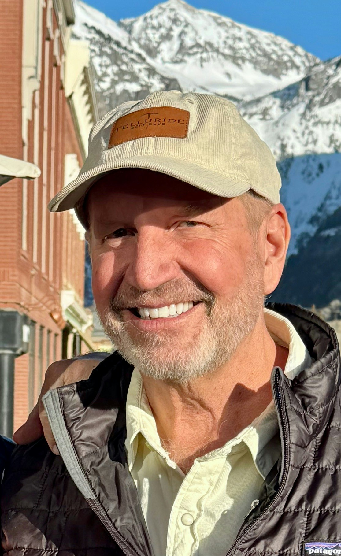 A person smiling in front of snowy mountains, wearing a cap and jacket, with a building in the background.