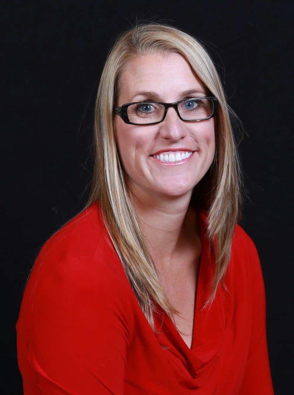 A person with long blonde hair, wearing glasses and a red top, smiling against a black background.