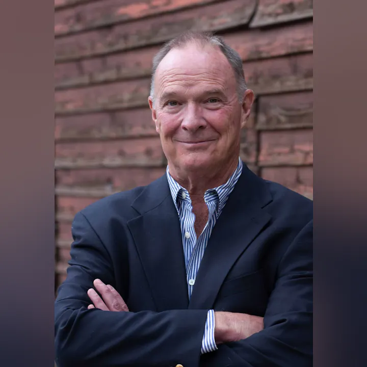 A person in a suit with arms crossed, in front of a wooden background.