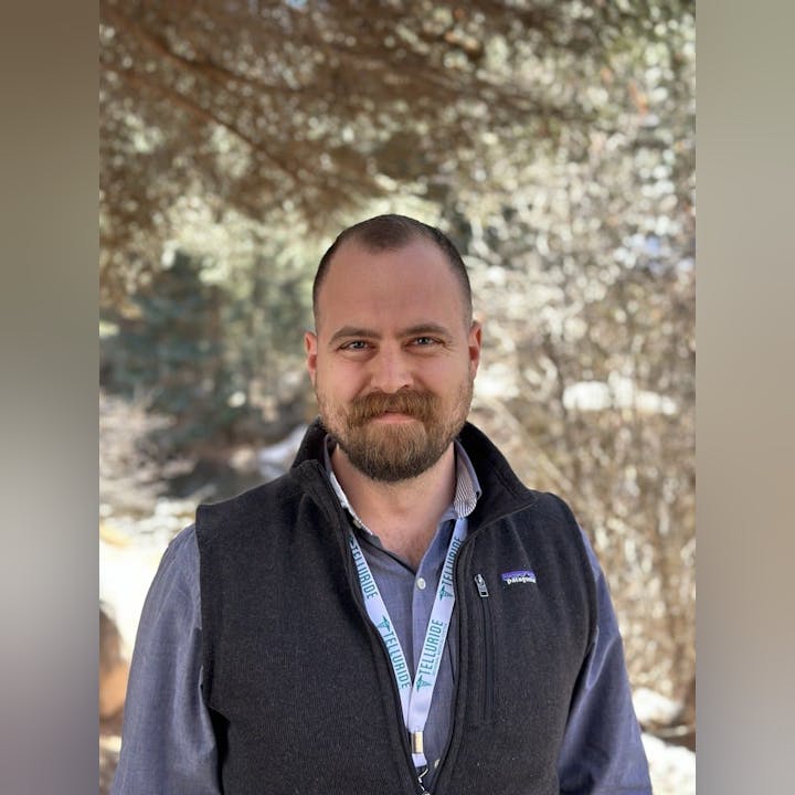 A man with a beard wearing a vest and a lanyard stands outdoors in front of trees.