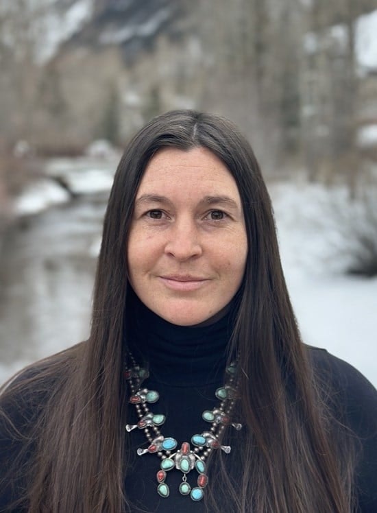 A person with long hair and a colorful necklace stands outside in a snowy setting, with a river and trees in the background.