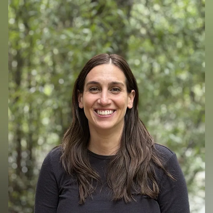 A smiling woman with long brown hair is posing outdoors amidst greenery.