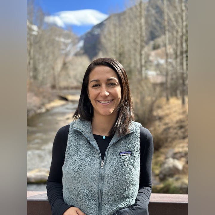 A person smiling outdoors, wearing a fleece vest, with a scenic background of a river and mountains.