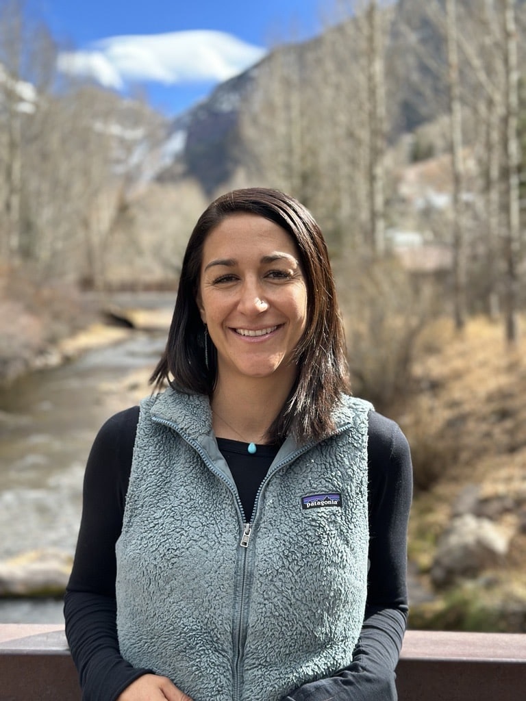 A person smiling outdoors, wearing a fleece vest, with a scenic background of a river and mountains.