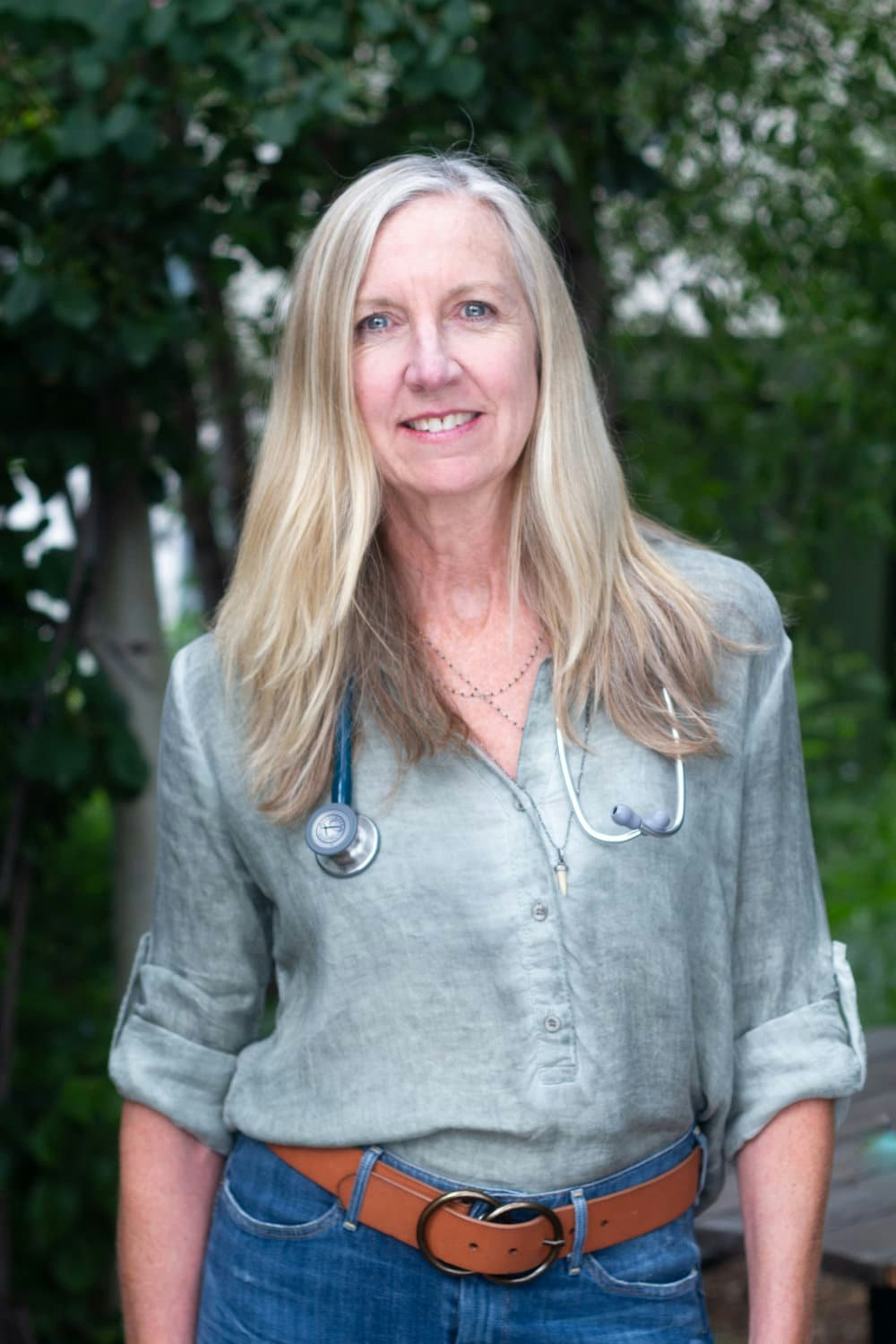 A woman with long hair, wearing a grey shirt, blue jeans, brown belt, and a stethoscope, standing outdoors.