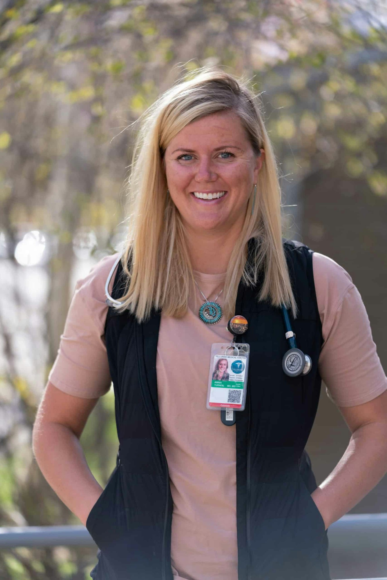 A person outdoors, wearing a medical ID badge and stethoscope draped on their shoulder, smiling at the camera.