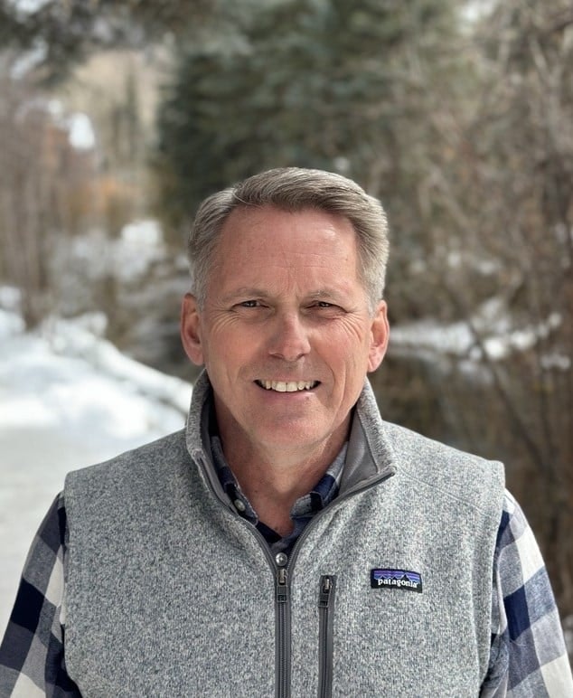 A person stands outdoors in a wintry setting, wearing a gray vest and plaid shirt with trees in the background.