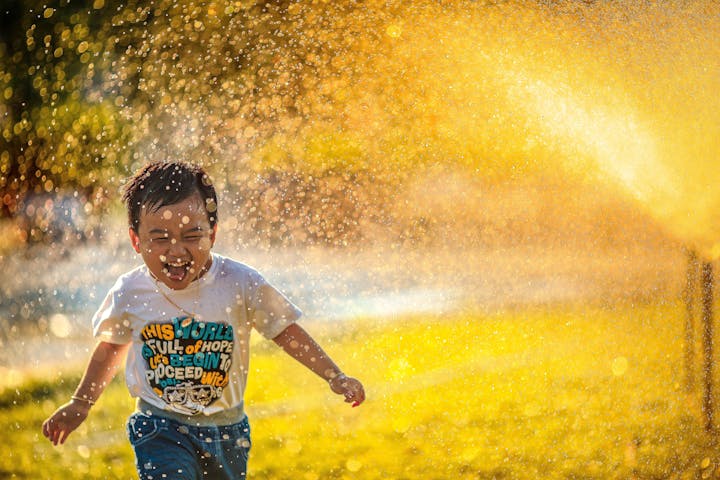 A joyful child runs through water spray, laughing and enjoying a sunny day, surrounded by sparkling droplets and a warm glow.