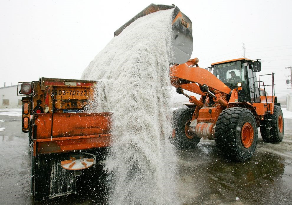 A front loader dumps salt into a snow plow truck during a snowfall.