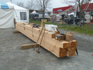 Large wooden beams are stacked together with tools and equipment on top, near a tent and fenced area.
