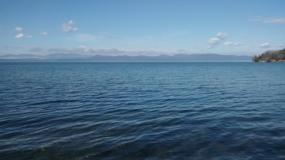 A calm lake with rippling water, distant mountains on the horizon, and a clear blue sky with a few clouds.