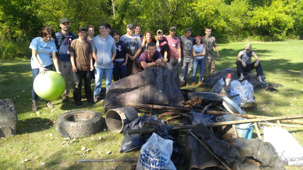 A group of people outdoors with collected trash and debris, including tires and bags, on the grass in a park or natural setting.