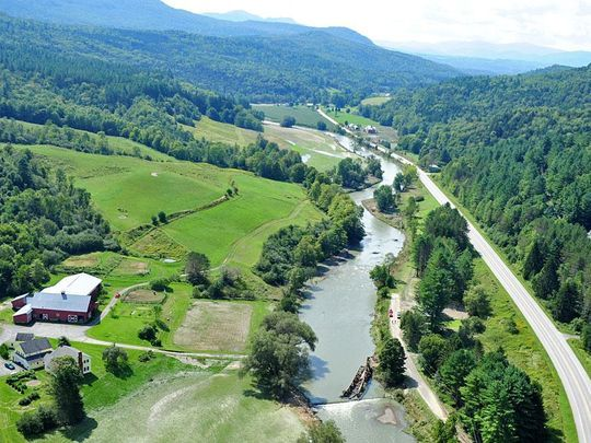 Aerial view of scenic countryside with a river, road, and houses nestled in lush green hills and trees.