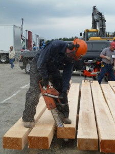A person is using a chainsaw to cut wooden beams at an outdoor work site, wearing safety gear including ear protection and a helmet.