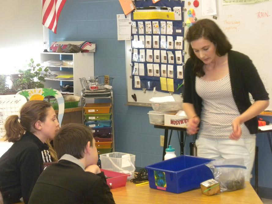 A teacher is engaged in a lesson with two students at a classroom table. There are educational materials and supplies around them.