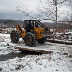 A yellow bulldozer with snow chains on the tires crosses a small wooden bridge in a snowy, wooded landscape.