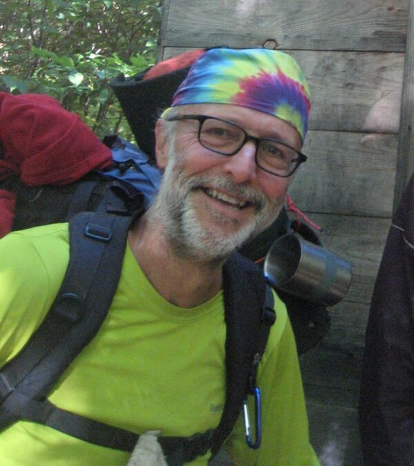 A smiling person with glasses, wearing a tie-dye bandana and hiking gear, including a backpack and a green shirt.