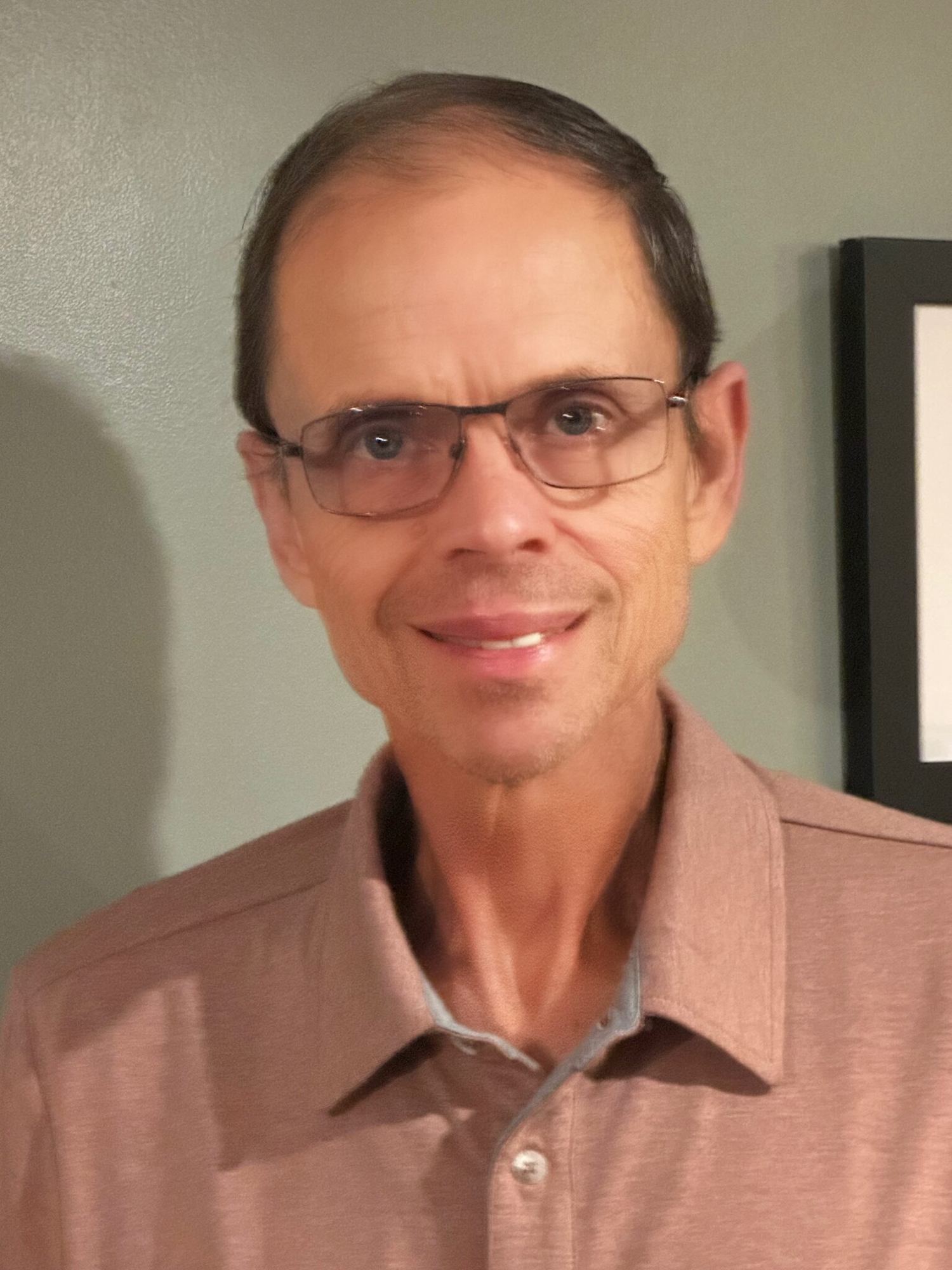 A person wearing glasses and a brown collared shirt smiles at the camera.