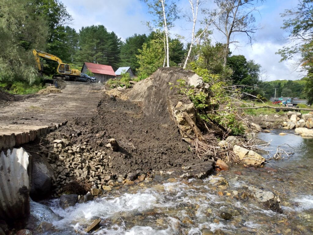 Excavator clearing a dirt path near a stream, with trees and buildings in the background.