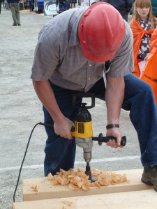 A person wearing a red hard hat uses a large drill on a wooden plank, with wood shavings scattered around.
