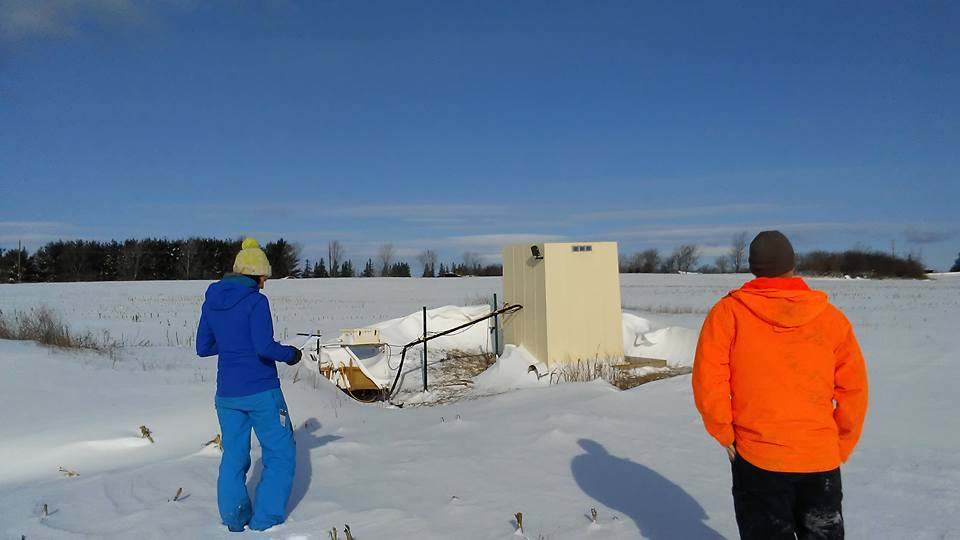 Two people in winter clothing looking at a structure in a snowy field.
