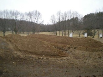 Rows of mulch or compost piles in a field with bare trees and overcast sky in the background.
