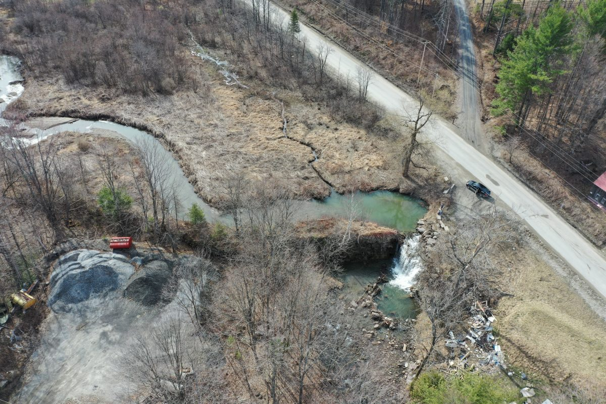 Aerial view of a rural area with a winding stream, a road, and a small waterfall. Sparse trees and a few scattered items are visible.