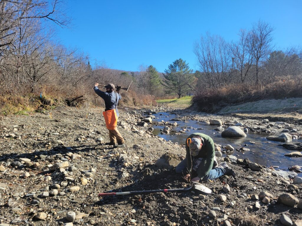 Two people working by a rocky stream; one is digging, and the other carries a tool. Leafless trees and clear blue sky in the background.