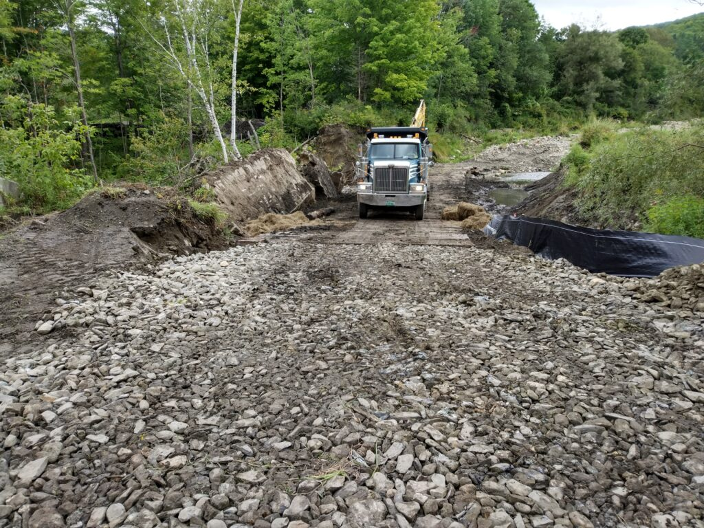 A construction truck on a rocky path surrounded by trees, with soil and tarp nearby.
