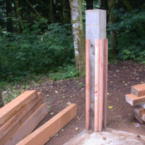 A concrete column with wooden planks attached, surrounded by more wooden beams and a forest background.