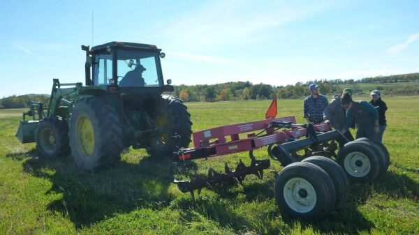A tractor pulling agricultural equipment in a field with three people standing nearby on a sunny day.