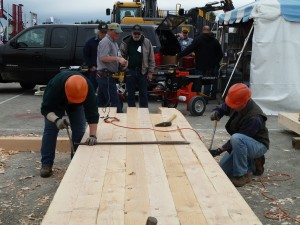 People working with large wooden beams at a construction site, wearing hard hats. A vehicle and equipment are in the background.