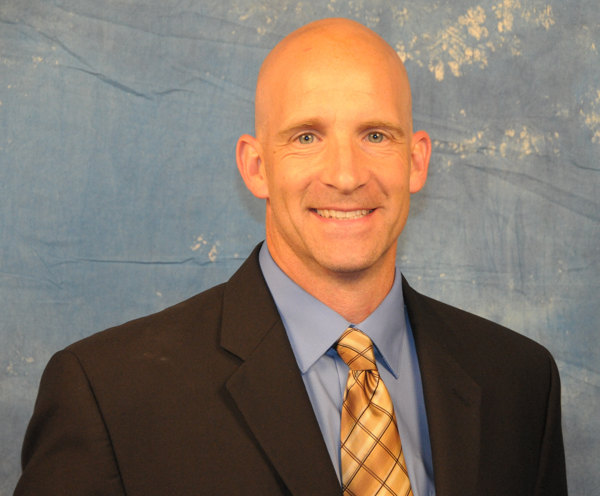 A bald man in a suit and tie smiling against a blue background.