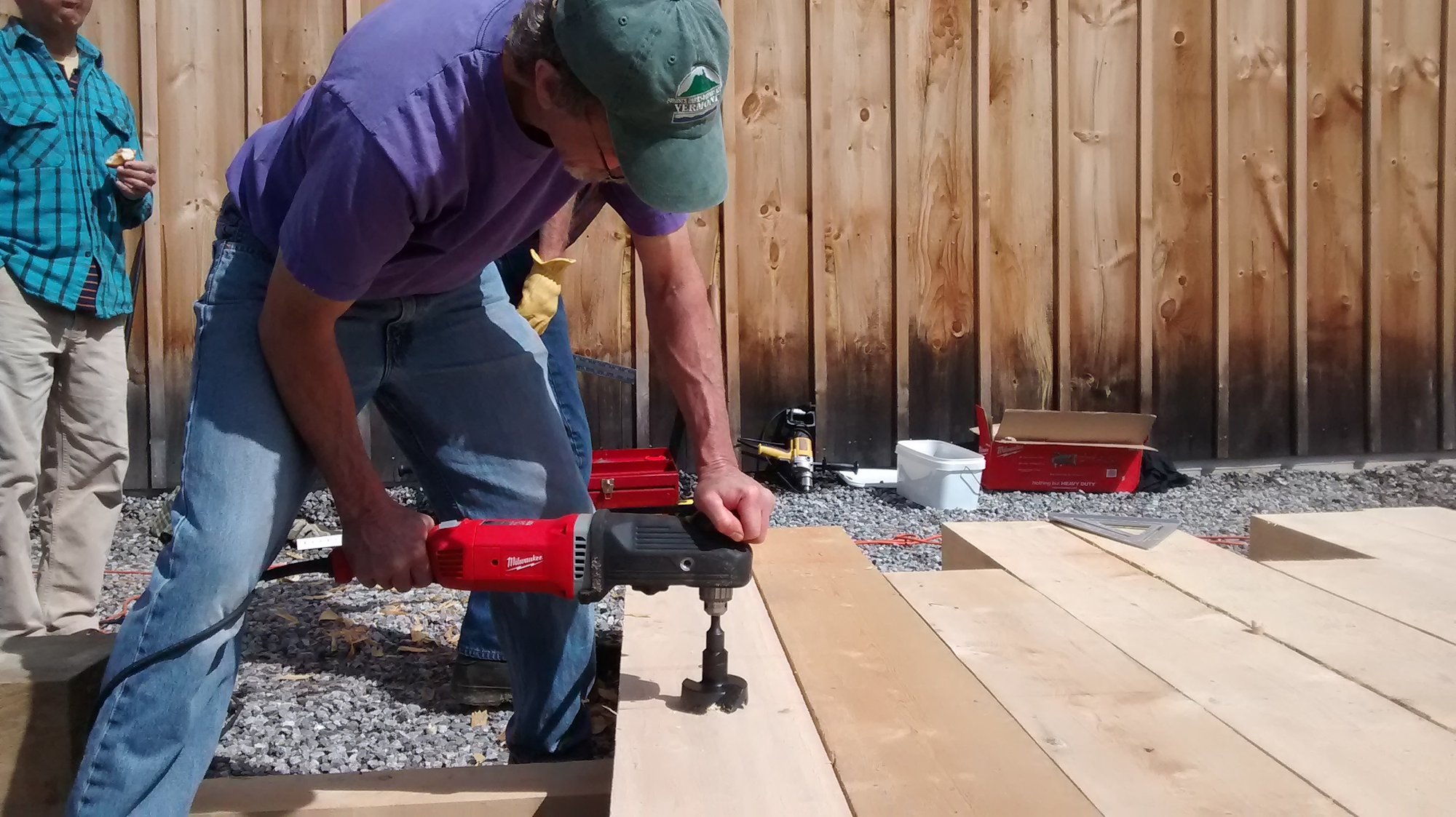 A person using a power tool on wood, with another person observing and eating. Tools and wood are scattered around.