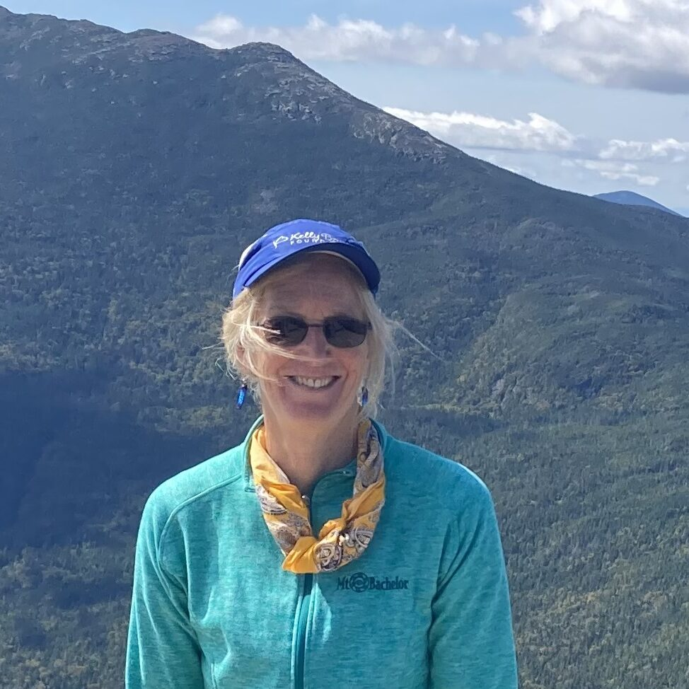 A person wearing a blue cap and sunglasses, smiling, with a mountain landscape in the background.