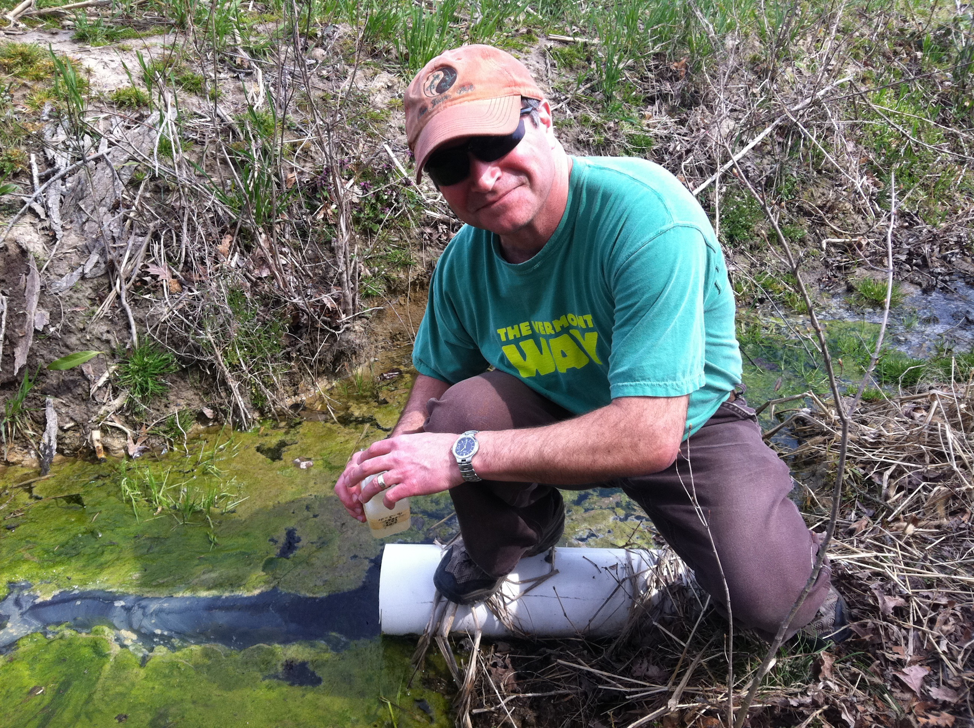 A person collecting a water sample from a stream, kneeling on a large pipe, surrounded by grass and foliage.