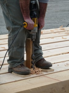 Person using a power drill on wooden beams, with wood shavings on the ground.